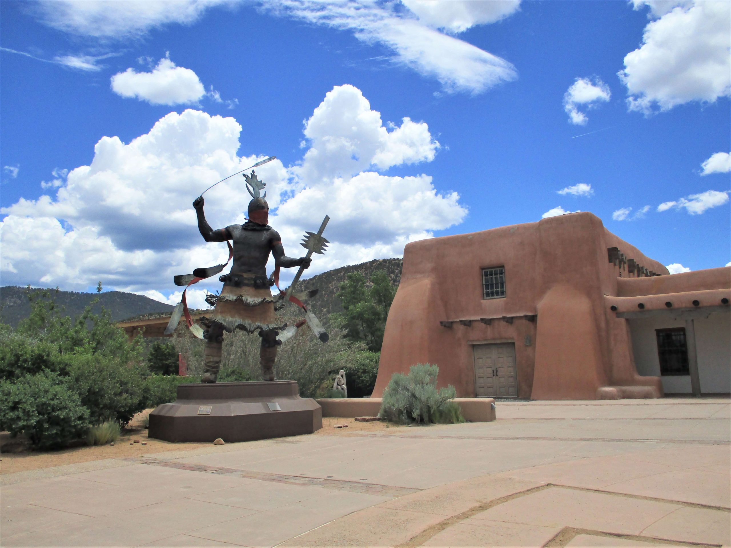 A bronze statue of a Native American dancer stands on a pedestal in an outdoor plaza. The figure wears traditional regalia, including a feathered headdress, fringed clothing, and a sash with curved metal ornaments. One arm is raised, holding a whip-like object, while the other grips a wooden staff. Behind the statue, an adobe-style building with earthy tones, small windows, and wooden beams blends into the desert landscape. The sky is bright blue with fluffy white clouds, and distant mountains add depth to the scene.