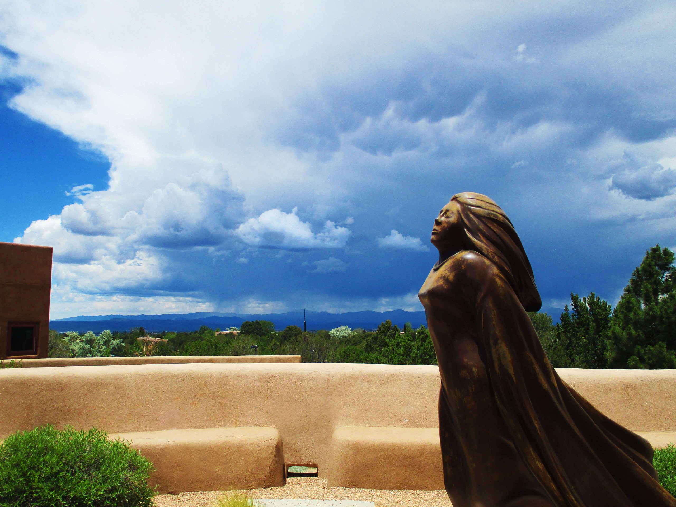 A bronze statue of a woman with long, flowing hair and a billowing cloak stands against a dramatic landscape. Her face is uplifted, as if feeling the wind. Behind her, a vast sky filled with billowing white and gray clouds stretches over distant mountains. The scene is framed by adobe-style architecture and desert greenery, evoking a Southwestern setting. (ChatGPT-generated)