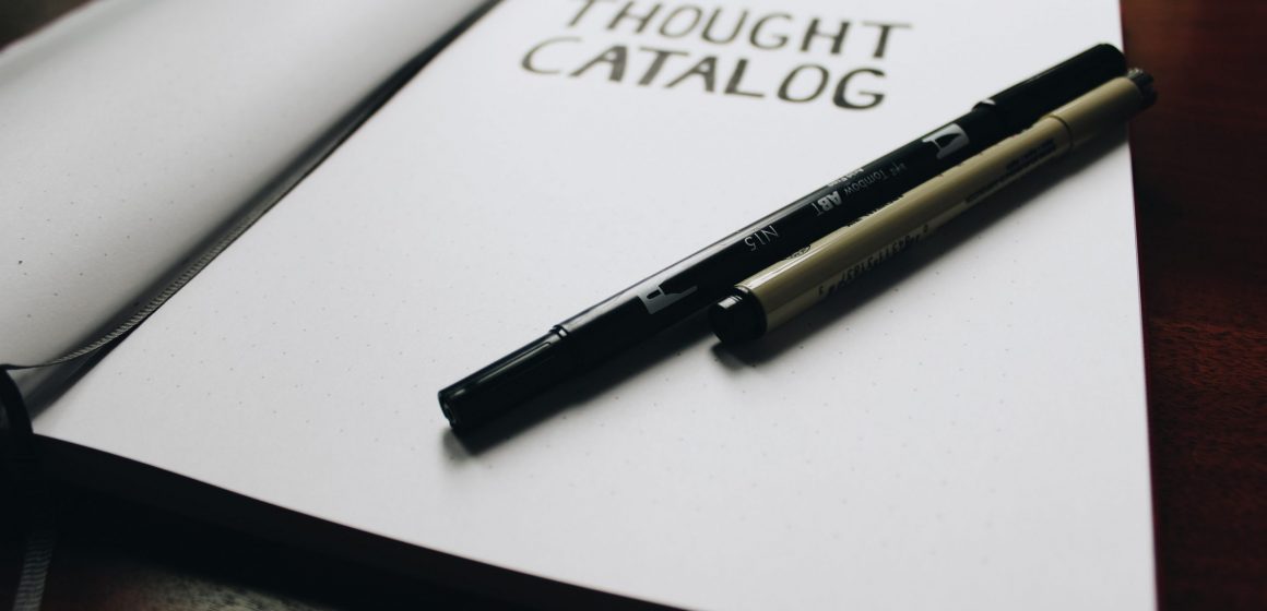 Open journal with text on it saying "Thought Catalog" with two pens laying down on a blank page ready for input.